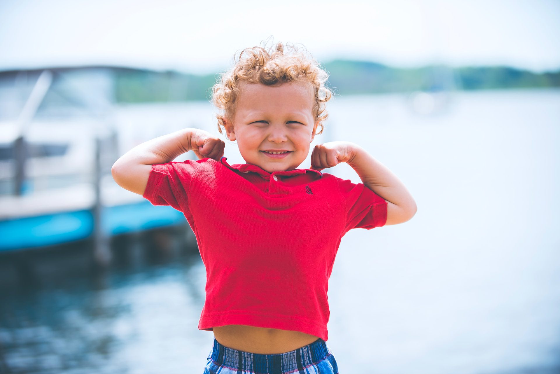 little boy in red shirt flexing his muscles in front of a lake